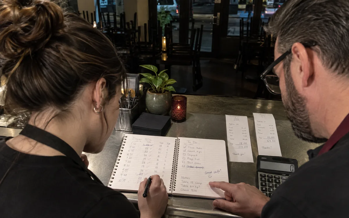 Scène illustrative : deux responsables consultent des reçus et un carnet à la fermeture du restaurant.