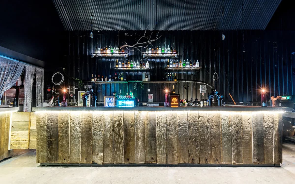 An empty wooden bar counter with illuminated bottle shelves in a dark interior.