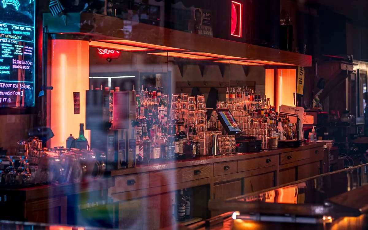 A warm-lit bar counter and stacked glasses seen through a reflective window.