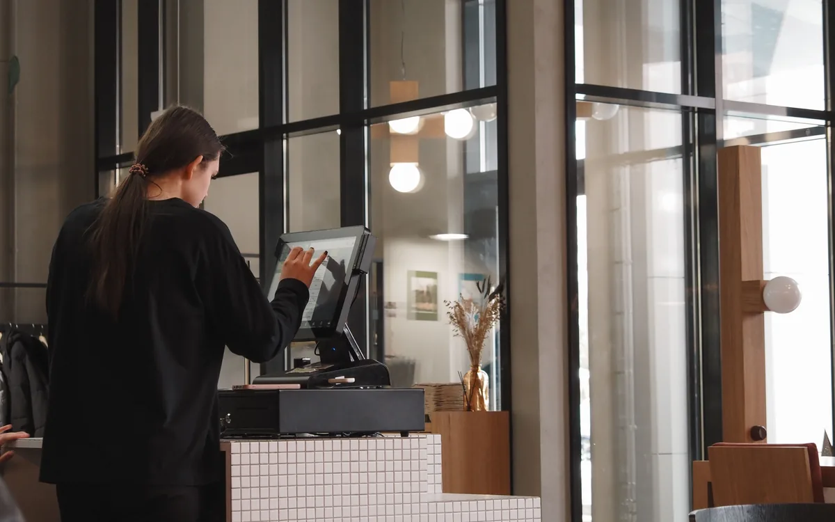 A café employee seen from behind using a touchscreen register beside tall windows.