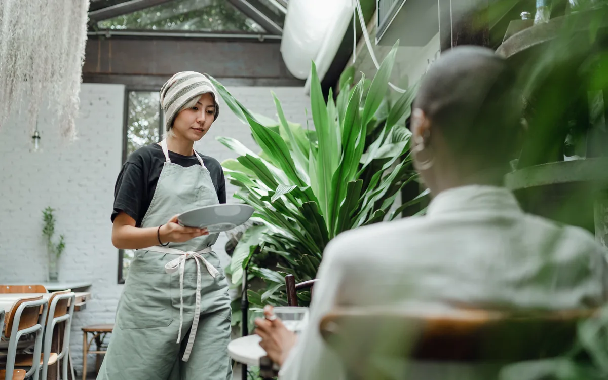 A café server carrying a plate to a seated guest beside green plants.